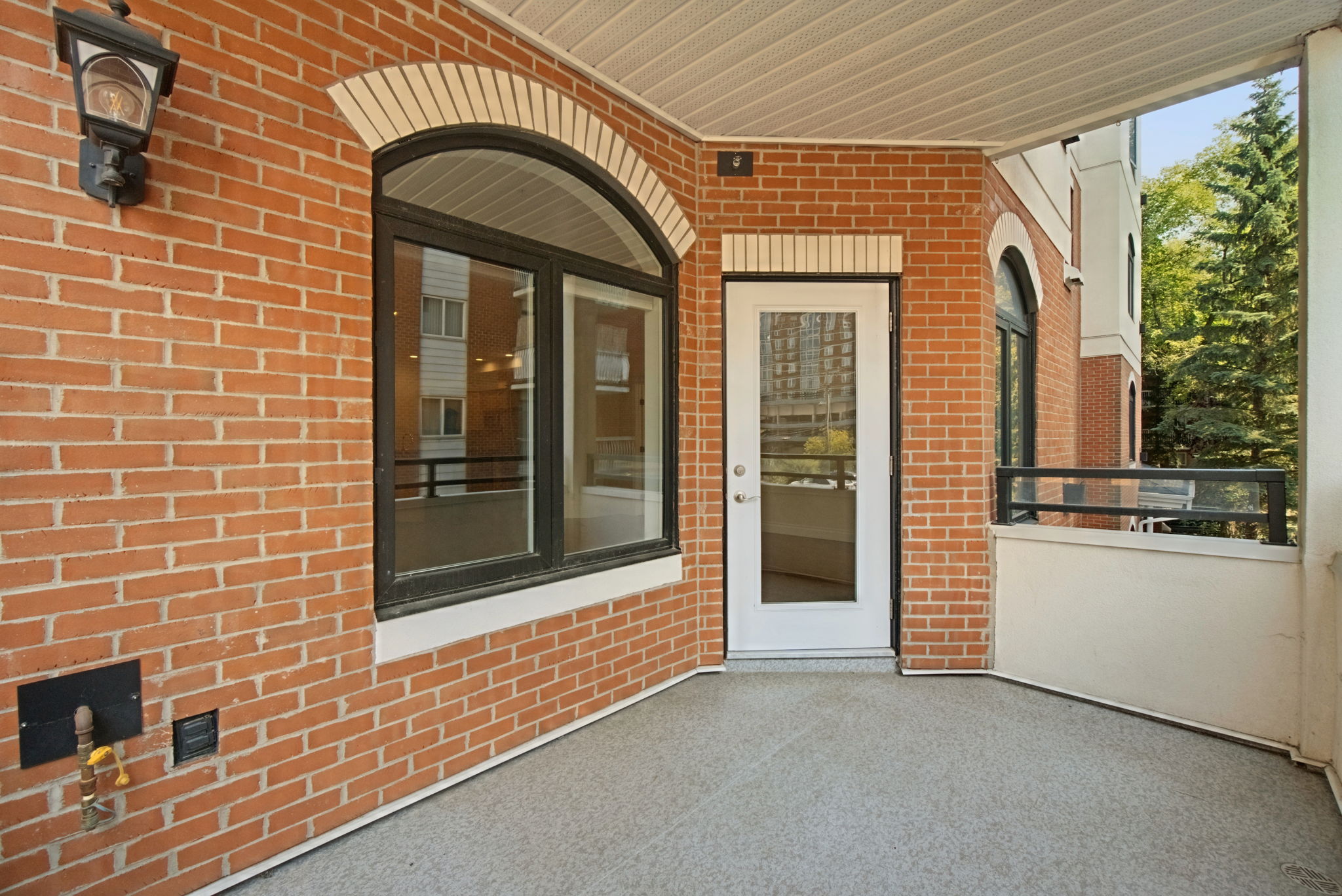 The covered balcony of unit 203, brick walls with an arched window and a glazed door into the living room, a light fixture on the wall, and the treed street beyond the railing.