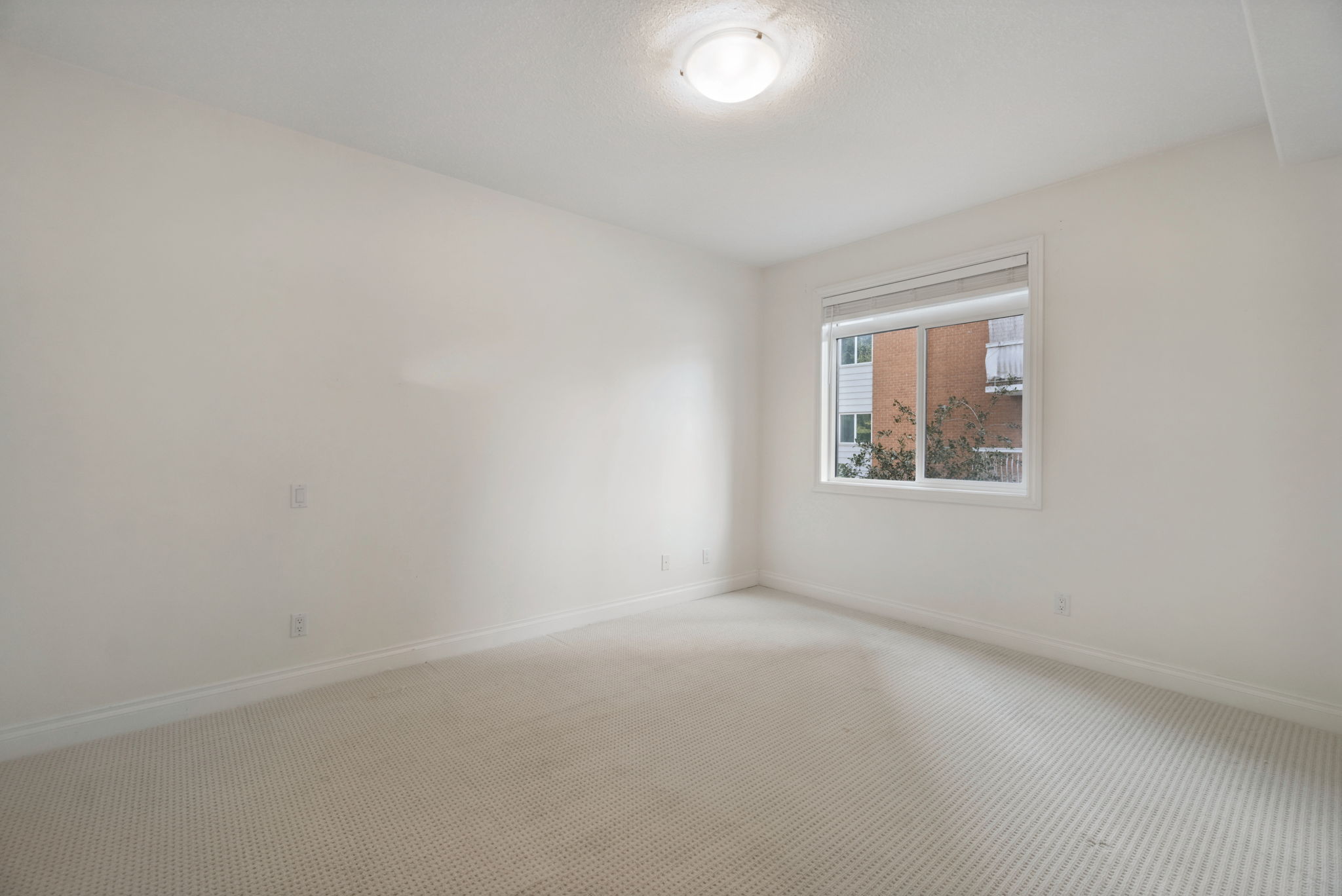 The second bedroom, carpeted, with a window looking out to the brick of the neighbouring building.