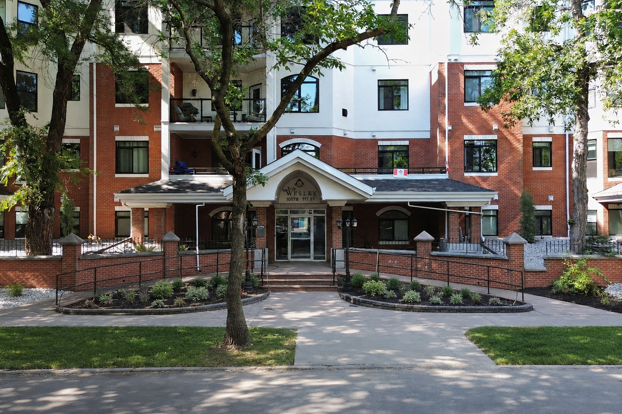 The front entrance of The Wesley, a gabled brick portico with the building's name over the door, framed by a mature elm and new plantings.