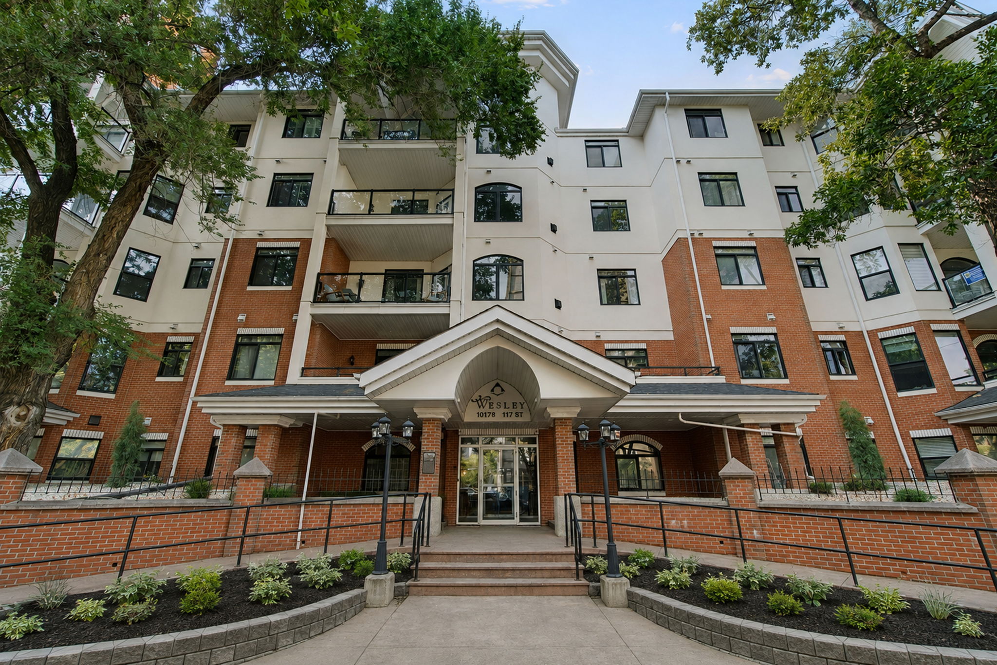 The front of The Wesley, a five storey brick and stucco building with covered balconies, seen through mature elms on 117 Street in summer.