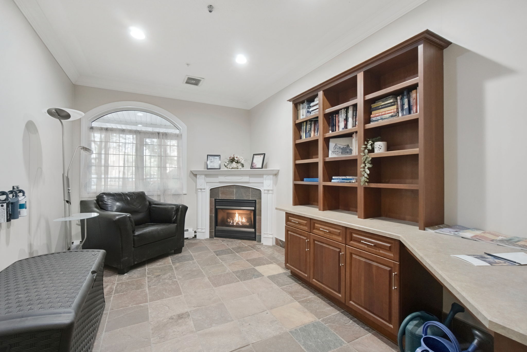 The library off the lobby, with a gas fireplace, armchairs, a bookcase and an arched window.