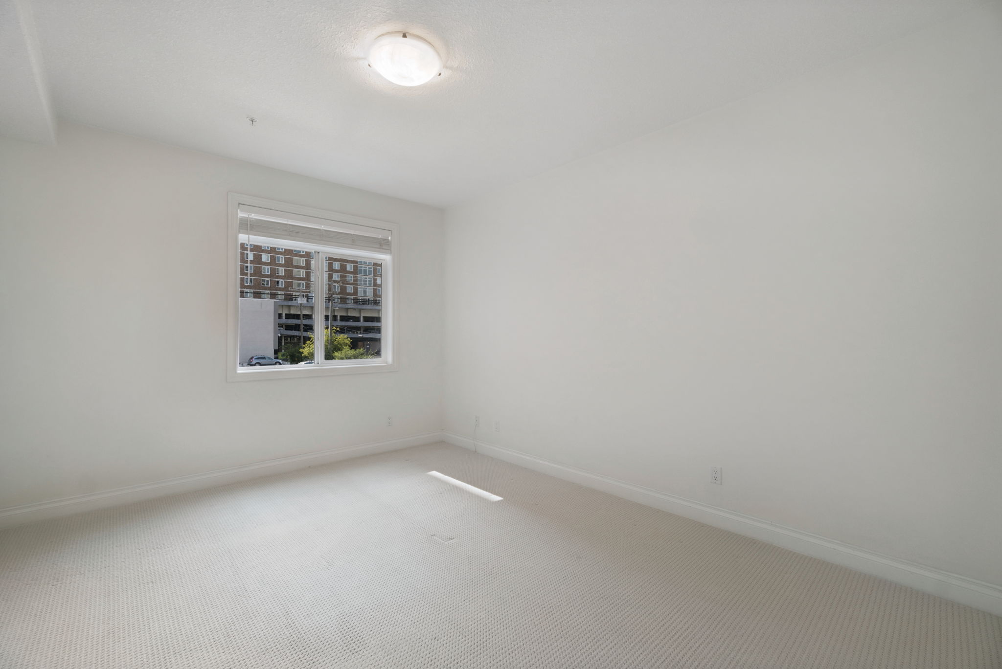 The primary bedroom, a long carpeted room with a window at the far end looking toward the neighbouring buildings.