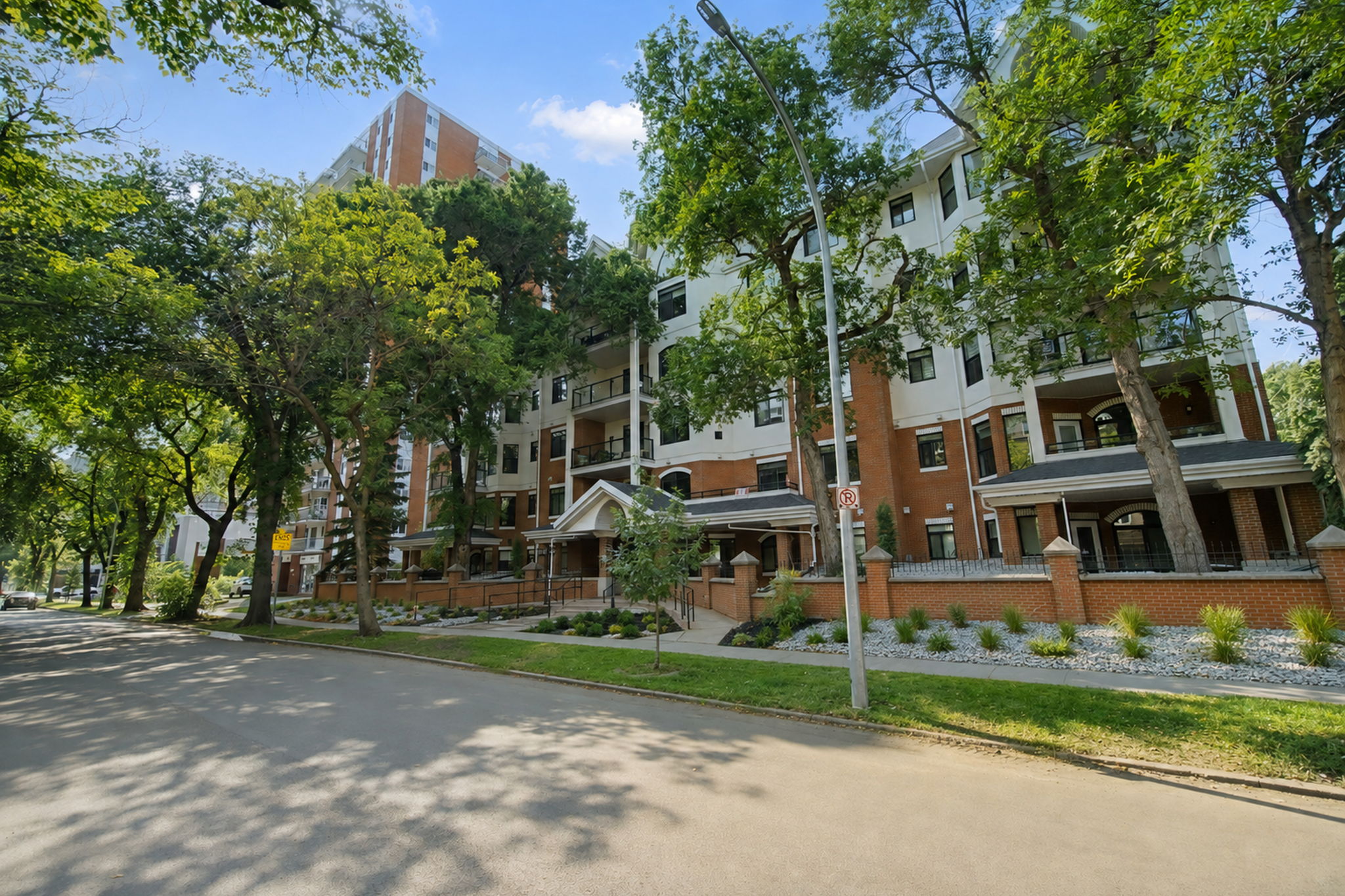 117 Street in summer, a wide sidewalk under mature elms with The Wesley's brick facade on the left.